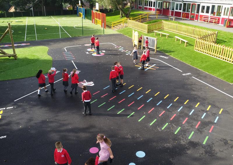 A playground with a variety of different playground markings installed on it. A line goes around the playground as a trail. A group of children are playing on the playground markings.